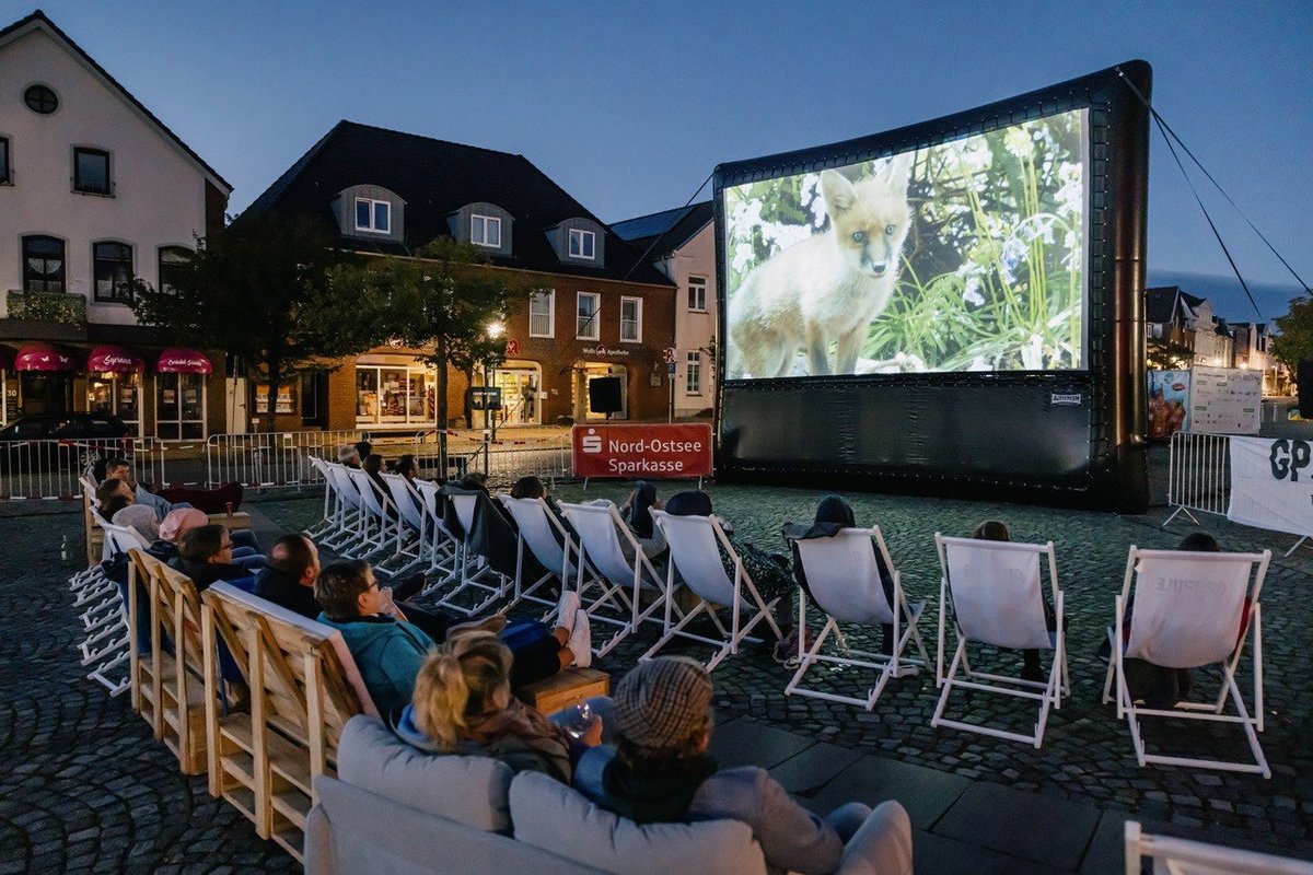 Das Open Air Kino auf dem Marktplatz sorgt für ganz besondere Momente. Foto: Nordseeküste Nordfriesland / Markus Rohrbacher Menschen sitzen auf Stühlen vor einer riesigen Kinoleinwand auf dem Marktplatz.
