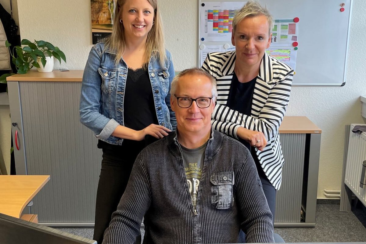Freude über den gelungenen Pilotversuch: Jessica Mühlenbeck, Amtsleitung Zentrale Dienste (rechts), Linda Jensen, Stellvertretende Amtsleitung Finanzen und Gunnar Maronde von der EDV-Abteilung des Amts Mittleres Nordfriesland. Foto: Felix Middendorf Zwei Frauen stehen nebeneinander hinter einem Sitzenden Mann.