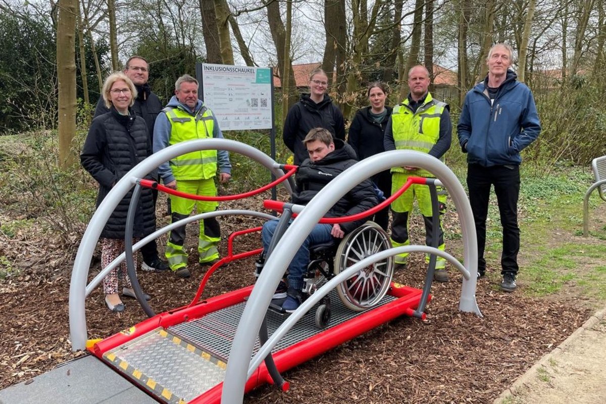 Ortstermin an der Inklusionsschaukel mit Bauhofmitarbeitern, Haus am Mühlenteich und Bürgermeister.  Eine Gruppe von Menschen steht um die Schaukel herum. Auf ihr befindet sich ein Mensch im Rollstuhl.